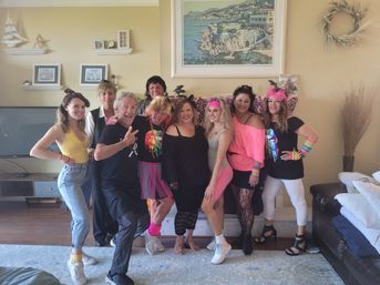 Smiling group of adults in colorful 1980s-style neon outfits and accessories posing for a themed party in a bright coastal-style living room with sofa, TV, and seaside wall art.
