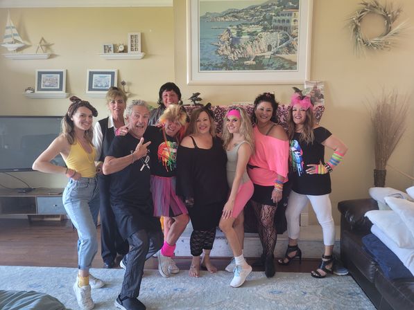 Group of adults in bright 1980s-inspired outfits and neon accessories posing and smiling in a sunny living room with coastal wall art, decorative shelves and a TV — playful retro party vibe.