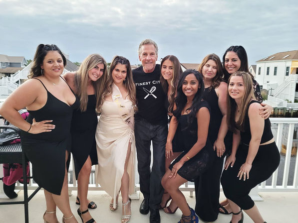Bachelorette party group photo on a waterfront balcony — bride-to-be in ivory dress and sash surrounded by friends in black dresses and a man, coastal canal houses in the background, overcast evening