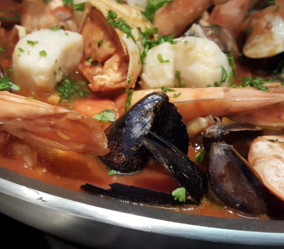 Close-up of a seafood stew in a stainless pan featuring prawns, mussels and scallops in a tomato broth garnished with chopped parsley