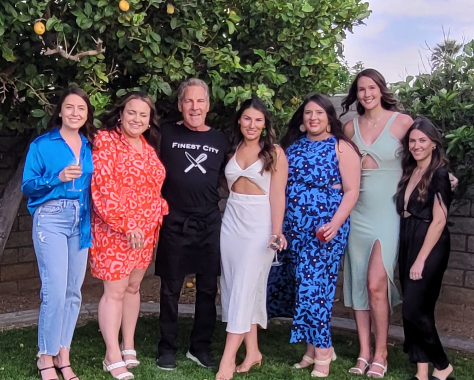 Seven adults smiling in a backyard garden under a lemon tree, wearing colorful summer dresses and casual outfits, posing close together and holding wine glasses.