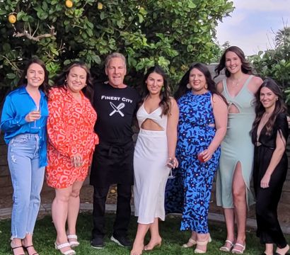 Seven friends posing on a backyard lawn under a lemon tree, wearing colorful semi-formal outfits and holding wine glasses at a summer garden gathering.