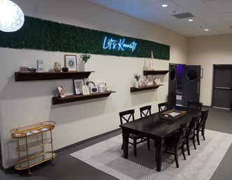 Modern coworking meeting area with a long dark wood table and eight chairs on a patterned rug, neon “Let’s Konnect” sign above a greenery accent strip, floating shelves with decor and a gold bar cart.