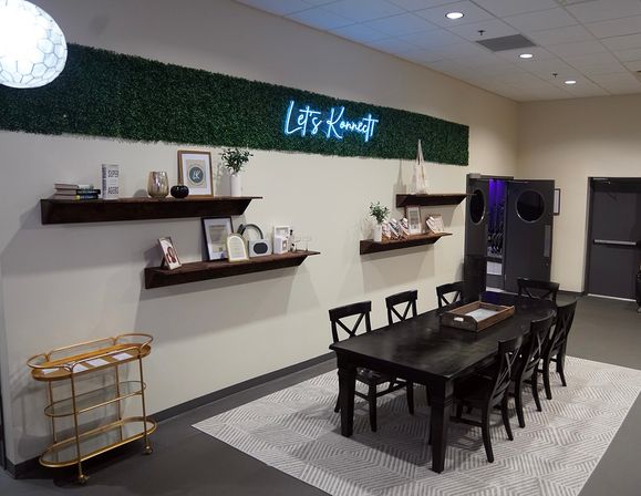 Modern coworking meeting area with a long dark wood table and eight chairs on a patterned rug, neon “Let’s Konnect” sign above a greenery accent strip, floating shelves with decor and a gold bar cart.