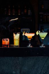Four vibrant cocktails lined up on a dark bar counter: a short berry-on-ice rocks drink, a tall yellow iced tropical with an edible flower, a chili-rimmed margarita with lemon and flower garnish, and a green martini with lime and olive — moody bar setting for craft cocktails and nightlife.