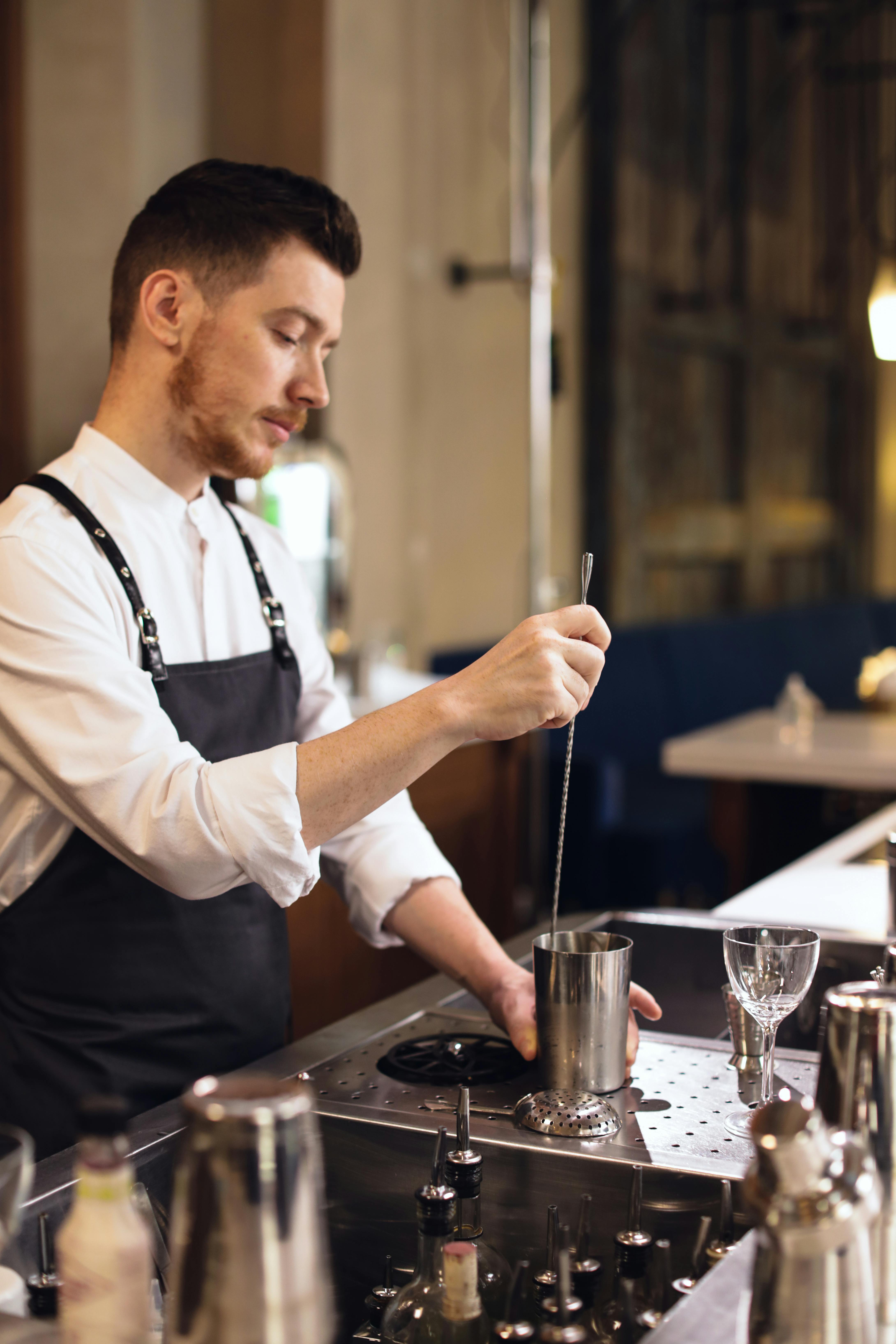 Mixologist stirring a cocktail in a metal shaker at an upscale cocktail bar, glassware and bottles on the polished counter.
