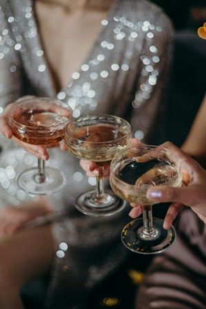 Three coupe glasses clinking with champagne at a festive party, hands raised over a sequined dress and twinkling bokeh lights