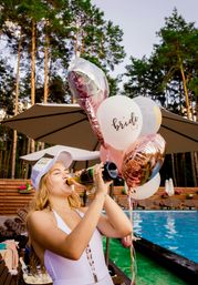Bride-to-be in a white swimsuit and 'BRIDE' cap chugs champagne poolside, holding pink and metallic 'bride' balloons at an outdoor bachelorette party with umbrellas and pine trees.