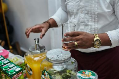 Close-up of a market vendor's hands holding a drinking glass above glass jars of citrus-and-mint infused water and orange juice, a gold wristwatch and ring visible.
