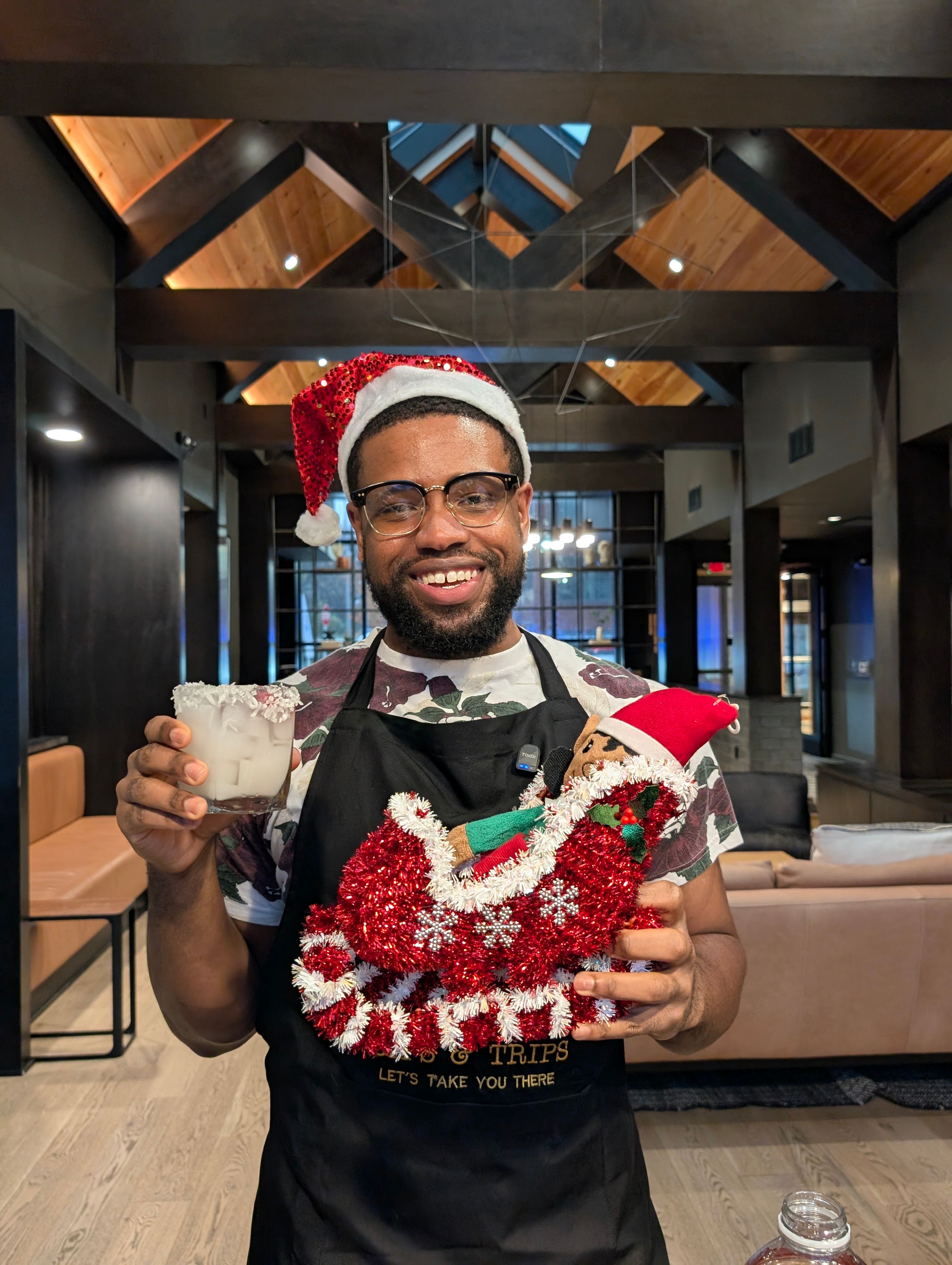 Smiling man in a Santa hat and apron holding a frosty holiday cocktail and a red tinsel sleigh with a small elf, standing in a cozy modern lobby with exposed wooden beams.