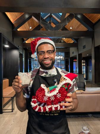Smiling man in a Santa hat and apron holding a frosty holiday cocktail and a red tinsel sleigh with a small elf, standing in a cozy modern lobby with exposed wooden beams.