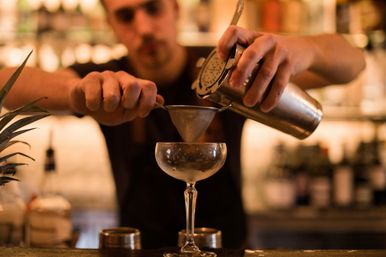 Bartender double-straining a craft cocktail from a metal shaker through a fine mesh strainer into a chilled coupe glass at a dimly lit cocktail bar