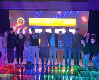 Smiling group posing on a colorful LED dance floor in front of a scoreboard reading Green Team 9186, celebrating a win at an indoor event.