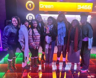 Group of eight women posing on a colorful LED arcade floor, one holding a trophy beneath a scoreboard reading "Green 3466" after a game-night win.