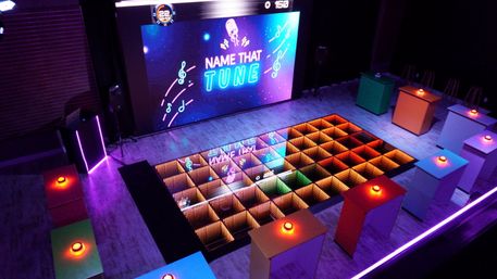 Indoor game-show stage with large LED screen reading 'Name That Tune', mirrored LED grid floor glowing rainbow colors, colorful contestant podiums with illuminated buzzers and neon purple ambient lighting.