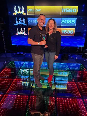 Two people smiling and holding a trophy on a glowing multicolored LED dance floor in an indoor game-show style arcade, with a large digital scoreboard behind showing 1st YELLOW 11560, 2nd GREEN 2085, 3rd ORANGE 1636.