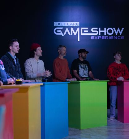 Group of contestants at a lively indoor game-show set, standing behind bright blue, green and pink podiums under neon stage lighting.