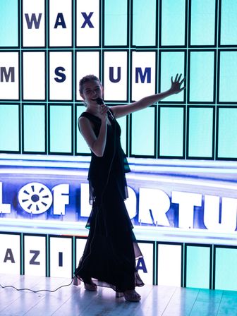Smiling woman in a black evening dress singing into a microphone on a brightly lit stage with a backlit letter grid, lively karaoke-style performance at an indoor event