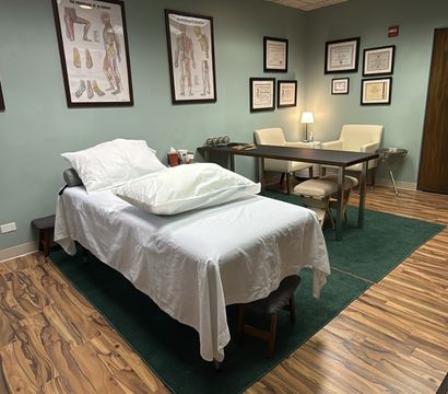 Cozy medical clinic exam room with a white-sheeted treatment table, anatomical charts and framed certificates on sage walls, a desk and chairs on a green rug, wood-look flooring and soft overhead lighting.
