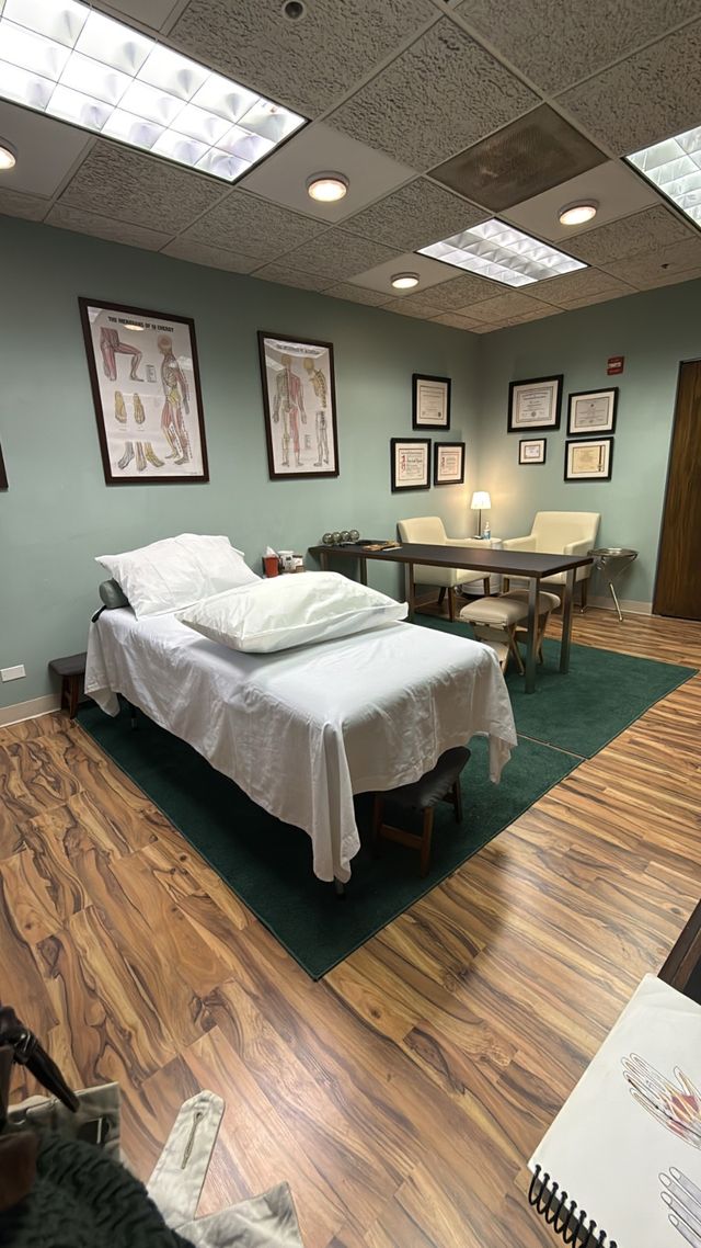 Cozy medical clinic exam room with a white-sheeted treatment table, anatomical charts and framed certificates on sage walls, a desk and chairs on a green rug, wood-look flooring and soft overhead lighting.