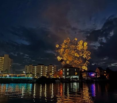 Night waterfront fireworks over a marina — sparkling golden bursts above lit condos and boats, colorful shoreline lights reflecting on calm water