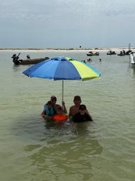 Family standing in shallow beach water under a blue-and-lime umbrella near a sandy sandbar, with boats and jet skis anchored offshore and an overcast sky