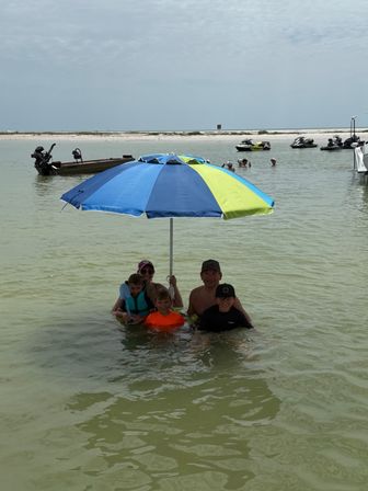 Family standing in shallow beach water under a blue-and-lime umbrella near a sandy sandbar, with boats and jet skis anchored offshore and an overcast sky