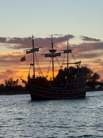Silhouette of a tall-masted pirate-style tour ship with passengers sailing on coastal waters at sunset, orange and purple sky reflecting on rippling sea