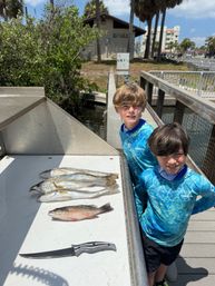 Two boys in matching blue fishing shirts on a sunny coastal dock beside a cleaning station displaying three spotted sea trout and a small snapper with a fillet knife.