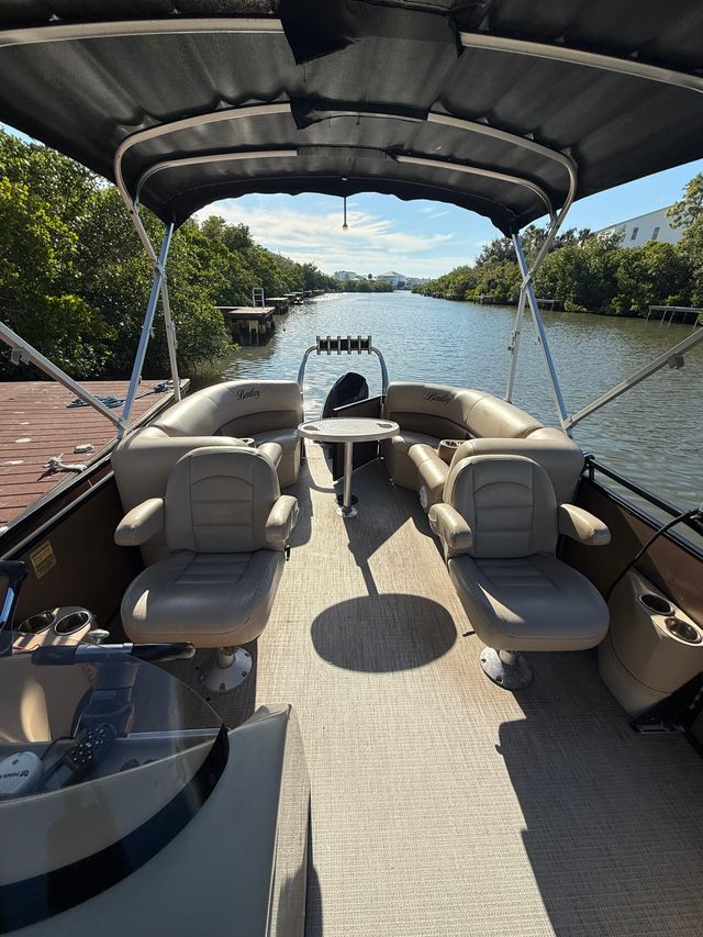 Sunlit pontoon boat interior with beige swivel seats and small round table under a black bimini top, docked along a calm mangrove-lined coastal canal with wooden docks and distant waterfront homes.