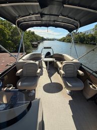 Pontoon boat interior with beige captain chairs and a round table under a black bimini, facing a calm mangrove-lined canal and docks on a sunny day — ready for a relaxing cruise.