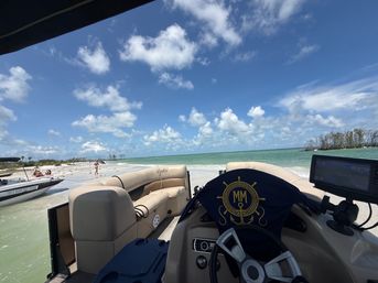 View from a pontoon boat helm toward a white sandy beach and turquoise water under a bright blue sky with puffy clouds, people wading on the sandbar and another small boat nearby.