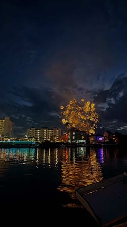 Sparkling golden fireworks over a waterfront marina at night, reflecting on calm harbor water with lit buildings and a boat in the foreground.