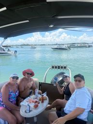 Four adults relaxing on a shaded pontoon boat with snacks and drinks, anchored in a calm turquoise bay with other boats and a bright blue sky