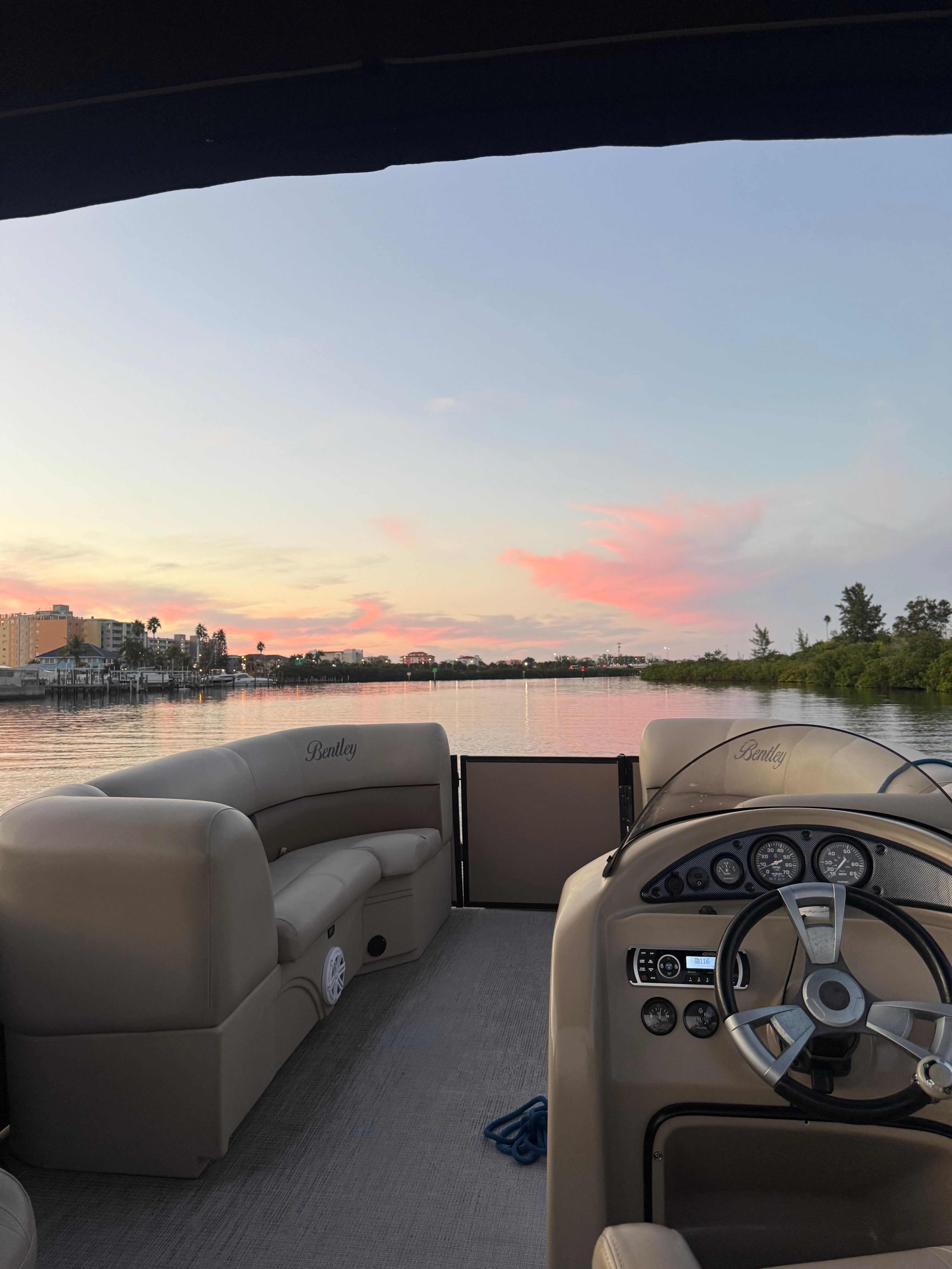 Sunset view from a pontoon boat: beige cushioned seats and steering console in the foreground, calm water reflecting pink clouds, and a distant shoreline with marina buildings and palm trees.