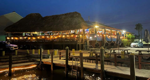 Tiki-style waterfront restaurant on wooden pilings at a marina, warm string lights and open-air seating, illuminated dock and picnic tables reflected in calm water at twilight