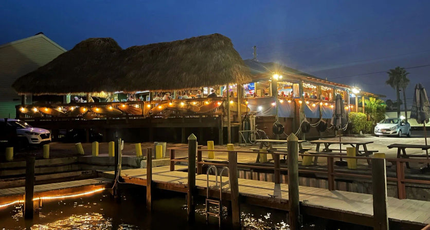 Tiki-style waterfront restaurant on wooden pilings at a marina, warm string lights and open-air seating, illuminated dock and picnic tables reflected in calm water at twilight