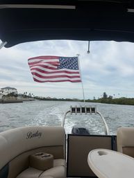 American flag flying from the stern of a pontoon boat cruising a coastal inlet, wake trailing behind, cushioned seats and table visible under an overcast sky.