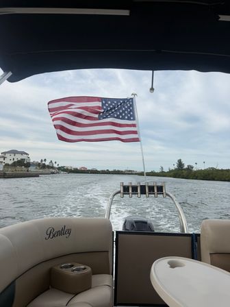 American flag flying from the stern of a pontoon boat cruising a coastal inlet, wake trailing behind, cushioned seats and table visible under an overcast sky.