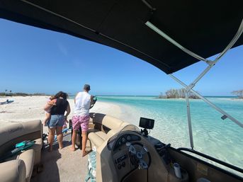Family on a pontoon boat beached at a white sand sandbar with clear turquoise water and bright blue sky — sunny tropical beach scene