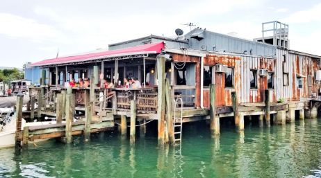 Rustic waterfront pier restaurant on wooden pilings with red awning and outdoor bar seating, diners overlooking emerald-green water and a small boat moored at the dock.
