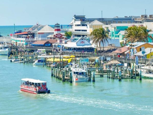Vibrant coastal marina and pier with colorful waterfront shops and palm trees, docks crowded with boats and a small red sightseeing ferry cruising turquoise water on a sunny day