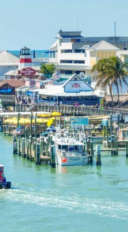 Vibrant coastal marina and pier with colorful waterfront shops and palm trees, docks crowded with boats and a small red sightseeing ferry cruising turquoise water on a sunny day
