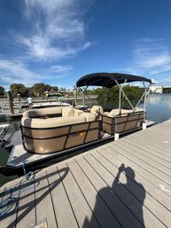 Tan pontoon boat with black bimini top moored at a sunny marina wooden dock over calm water under a vivid blue sky, ready for a leisure cruise.
