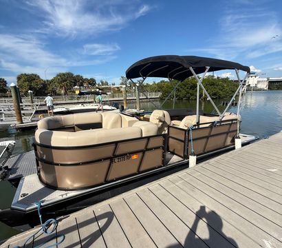 Tan pontoon boat with black bimini top moored at a sunny marina wooden dock over calm water under a vivid blue sky, ready for a leisure cruise.