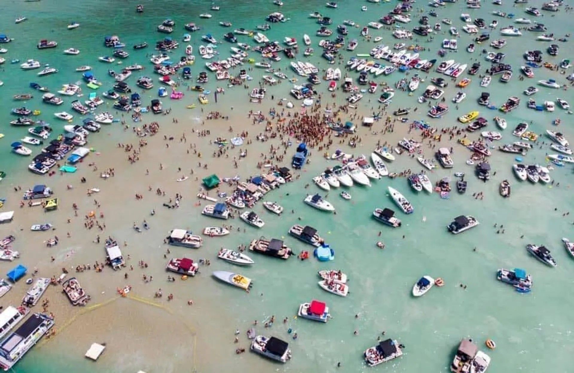 Aerial view of a lively sandbar boat party with dozens of anchored boats in clear turquoise shallow water and hundreds of people gathered on the sand and swimming.