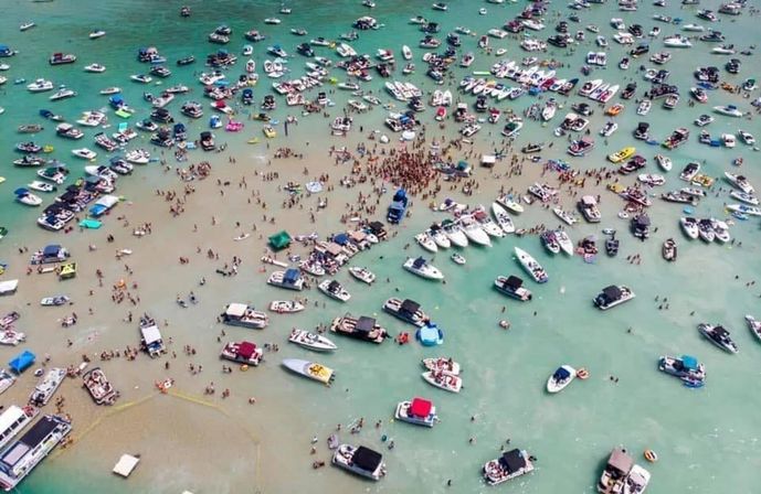 Aerial view of a lively sandbar boat party with dozens of anchored boats in clear turquoise shallow water and hundreds of people gathered on the sand and swimming.