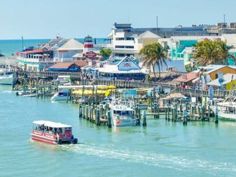 Vibrant coastal marina with colorful waterfront shops and restaurants, palm trees along a bustling pier, docked boats and a red tour boat cruising turquoise water.