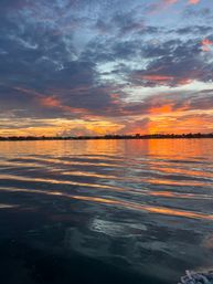 Vibrant orange and purple sunset over a calm coastal bay, dramatic clouds and glowing ripples reflecting light on the water with a silhouetted shoreline