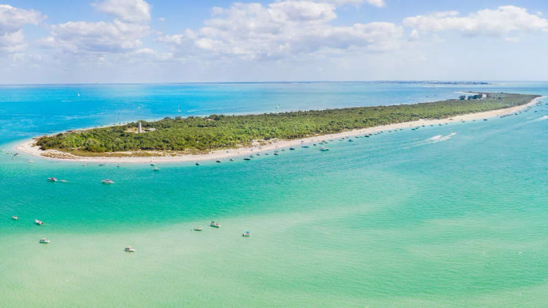 Aerial view of a long, green barrier island with white sandy beaches and a small coastal tower, surrounded by turquoise waters dotted with anchored boats under a sunny blue sky